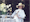 Film still of a woman wearing a yellow wide-brimmed sun hat and a white dress, with a dog on her lap. She is smiling and is next to a table with a vase daffodils on top.