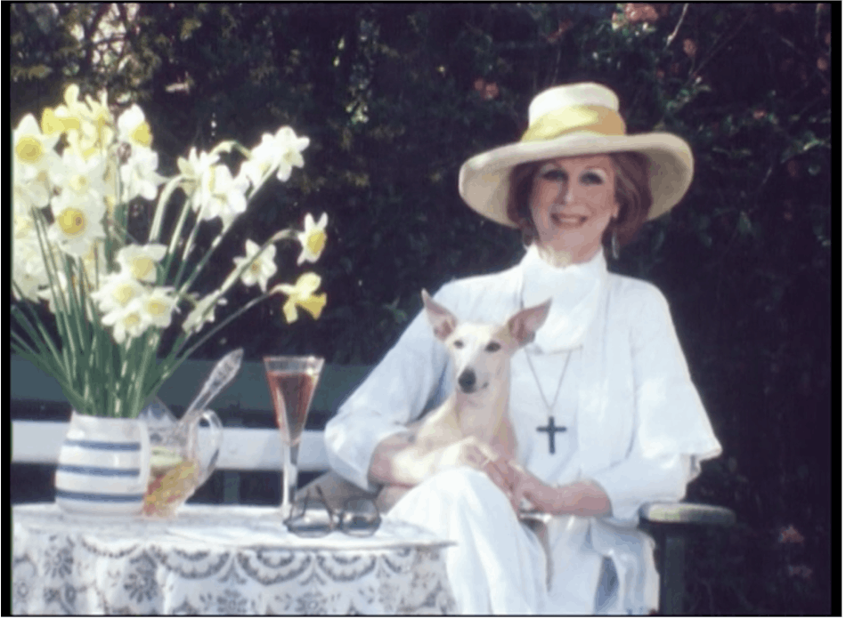 Film still of a woman wearing a yellow wide-brimmed sun hat and a white dress, with a dog on her lap. She is smiling and is next to a table with a vase daffodils on top.