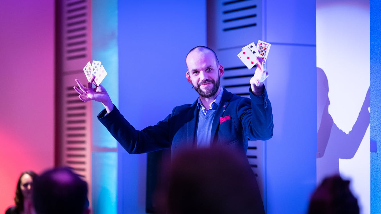 Photograph of a male magician holding up 2 playing cards in each hand, looking directly to camera. In the foreground are the backs of the heads of his audience. The magician is bathed in the pink and blue of the stage lighting.