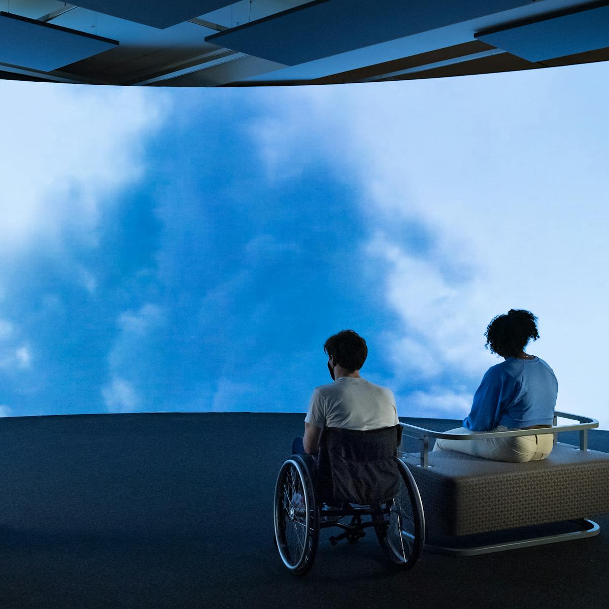 Colour photograph of three people watching a very large, curved screen. The young man on the left is standing up with his hands behind his back. The young man in the middle is in a wheelchair, to the right of a girl who is sat on a chair. On the screen is a scene showing blue sky and white clouds.