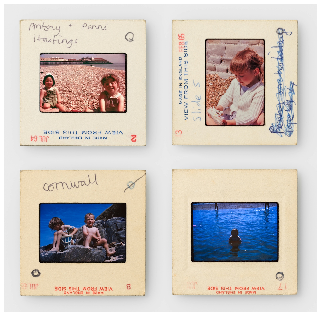 Photograph of four colour 35mm transparencies in a 2 by 2 grid, each mounted in a cardboard side holder, all resting on a white background. The transparencies on the top row show a young girl and her brother on holiday sitting on a shingle beach. In one image there is a pier in the background. The slide mounts have the words 'made in England. View from this side' printed on them. Hand written notes on the mounts read, "Antony and Penni Hastings" and "Slide 5". The transparencies on the bottom row show the same siblings but on a different beach with large rocks and a deep blue coloured sea. The slide mounts have the words 'made in England. View from this side' printed on them. A hand written note on one of the mounts reads, "Cornwall".