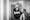 Black and white photograph of an individual at home in the process of hoisting themselves from their wheelchair.