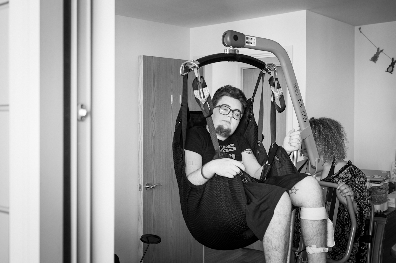 Black and white photograph of an individual at home in the process of hoisting themselves from their wheelchair.