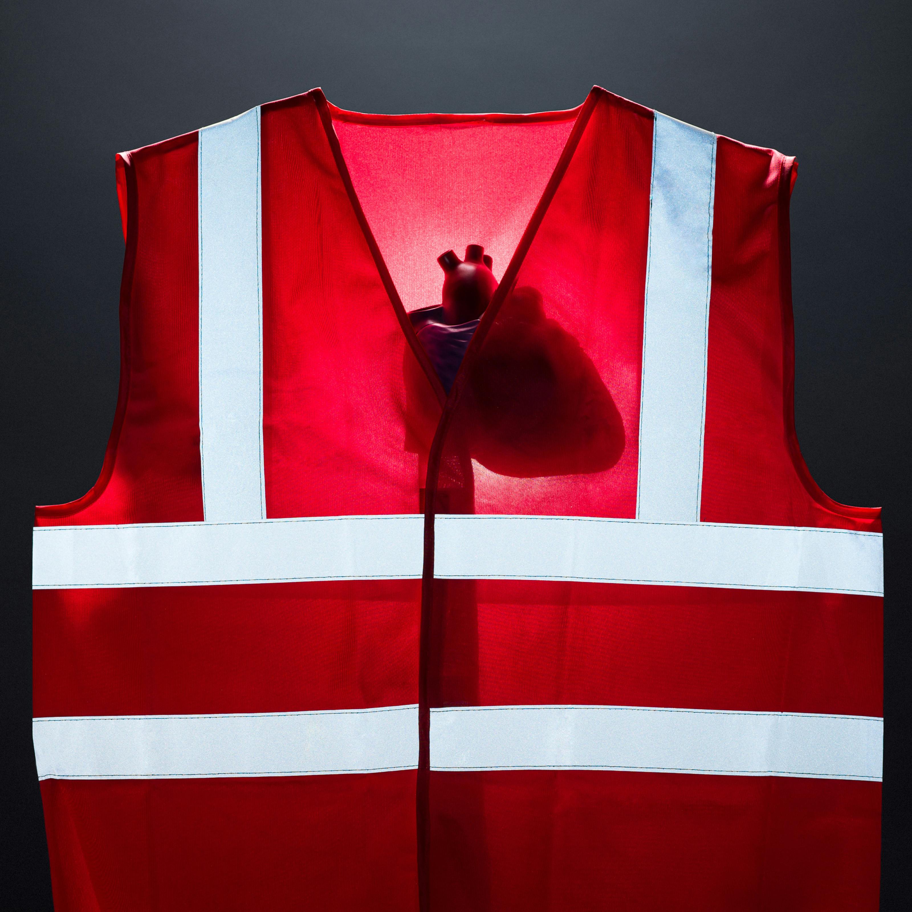 Photograph of a bright red high visibility vest against a dark grey background. The vest is lit from behind revealing the silhouette of a model of a human heart. The reflective strips on the front of the vest are bright with white light.