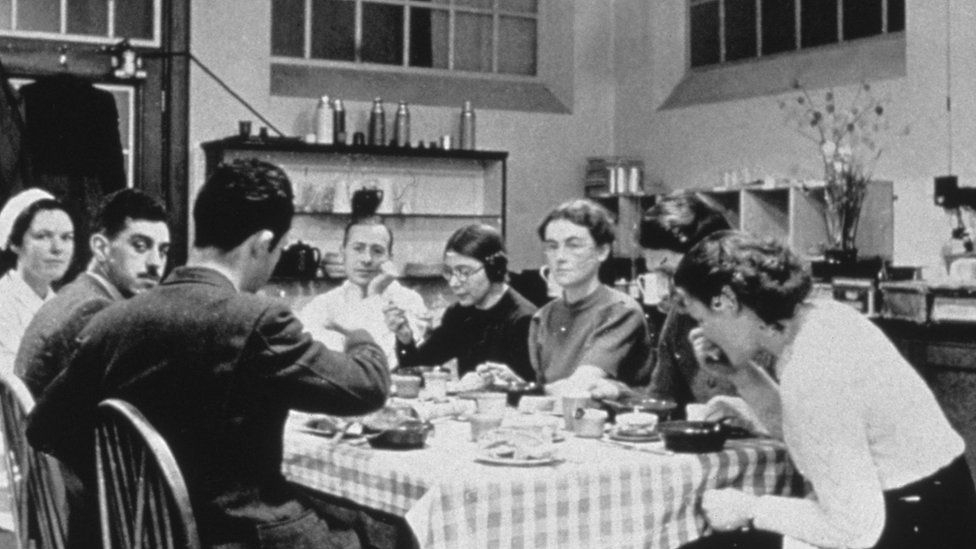 Black and white photograph showing people sitting and eating around a kitchen table with a gingham tablecloth. In the centre is a woman wearing a black shirt, bringing a spoon to her mouth: this is Elsie Widdowson. To her right is a man with a white shirt, his chin resting on his hand, and this it Robert McCance. 