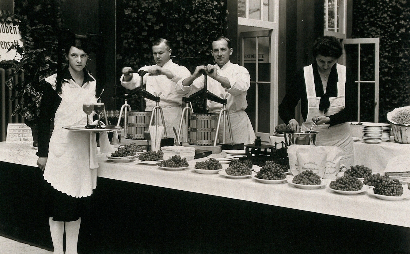 Photograph of a table laid out with bunches of grapes on plates. Behind it stand three servers, two of whom are pressing grapes; in front of it stands a server with two glasses of grape juice on a tray.
