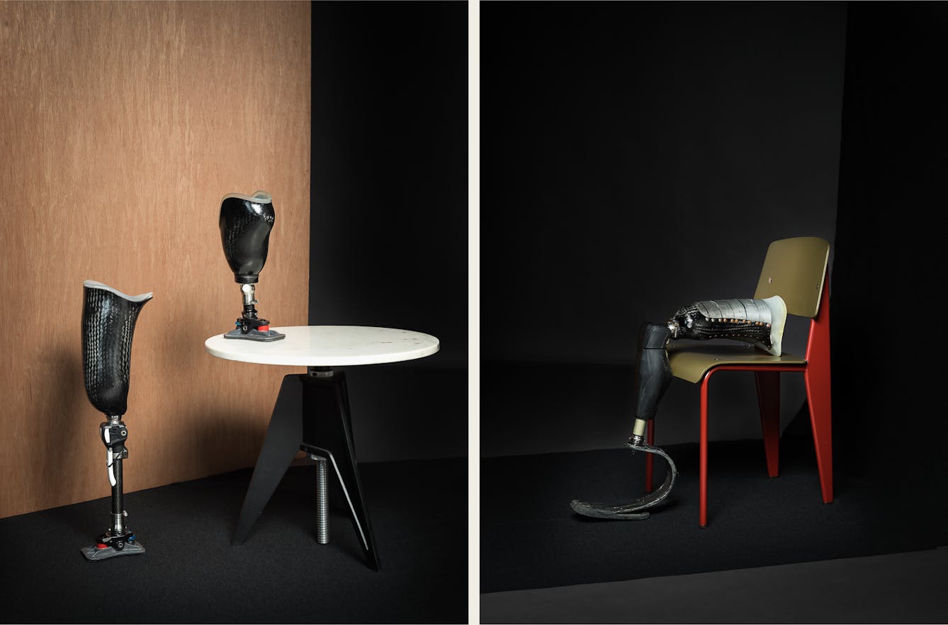 Diptych. The photograph on the left shows a table with a white top and a black base, resting on a black floor. There is a wooden panel behind the table. On top of the table there is a prosthetic limb and on the floor, there is another prosthetic limb standing up. 

The photograph on the right shows a black floor and backdrop. There is a chair with a red base and legs and a green seat. On the chair, a prosthetic limb is resting, with the base of it touching the ground. 