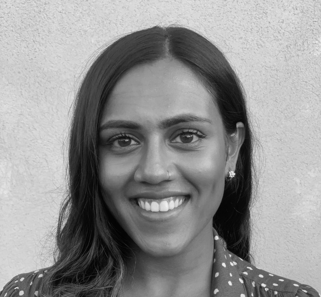 Black and white photograph of the head and shoulders of a young South Asian woman with long black hair. She is smiling and looking directly at the camera.