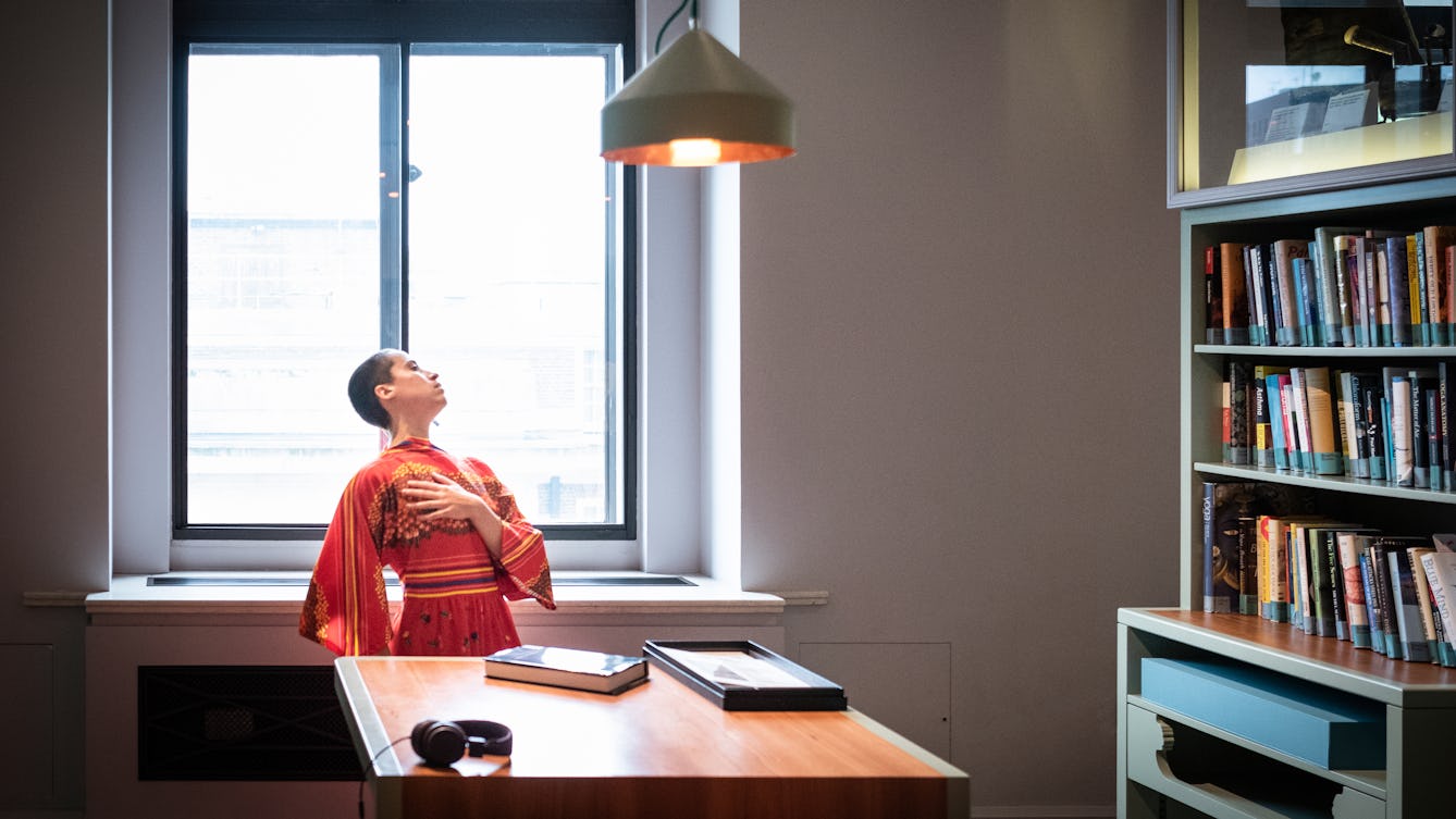 Photograph of a museum gallery environment with tables, a bookshelf full of books and a large window. In front of the window a performer with short cropped hair and red and yellow flowing dress, is mid performance, clutching their hand to their chest, their neck and head stretched back. Resting on the tabletop is a book and a set of headphones. Above the table is a copper lampshade painted green just appearing into the frame.