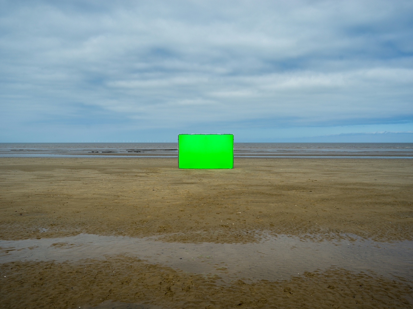 Colour landscape photograph showing a beach scene. The scene is split in half horizontally, with a blue sky and cloud covered upper and a sandy beach lower, with a thin strip of sea and breaking waves across the middle. In the centre of the image, some way off is a large rectangular photographic background frame standing up vertically. Stretched across the frame is a vibrant green chroma key fabric. In the foreground running across the sand from left to right is a thin ribbon of water in the sand.