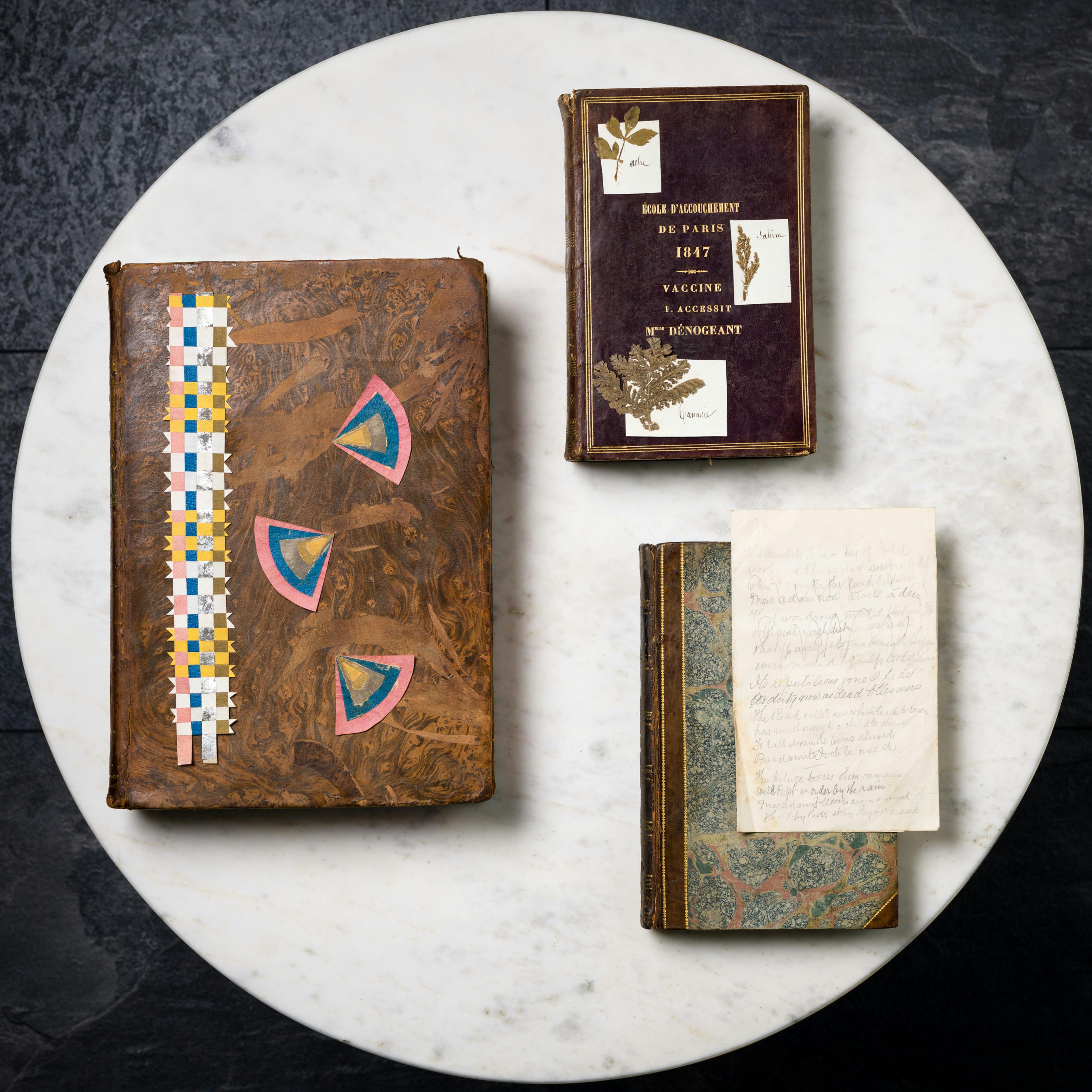 Three old books displayed on a marble tabletop, each with items on top. The first has some paper jewellery, the second has three pressed plants with name labels, and the final one has a sheet with pencil verse scribbled on it.