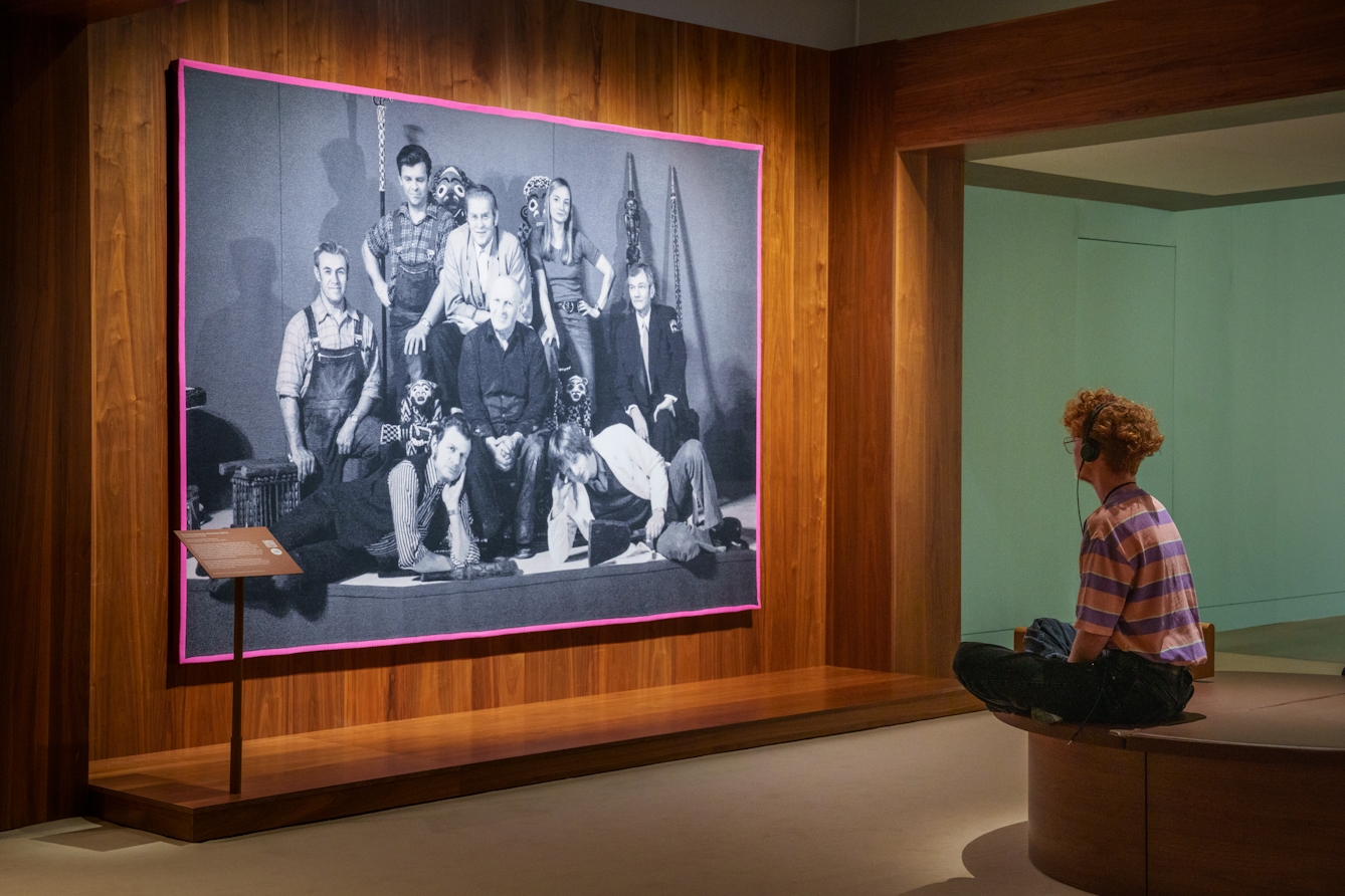 Photograph of a gallery exhibition space showing large tapestry hung on a wooden wall. The tapestry is made up of a black and white, archive photograph, from the late 20th century showing a group of white men and women gathered in a group within an exhibition exhibit. One of them is seated on an ornate throne with the others perched and lying around him, some holding museum artefacts in their hands. The tapestry is framed with a bright pink border. Surrounding the tapestry is a wider wooden structure containing seating and alcoves. Sat crosslegged on a seat in front of the tapestry is a gallery visitor in a striped top. They are listening to the gallery audio guide on a pair of headphones.