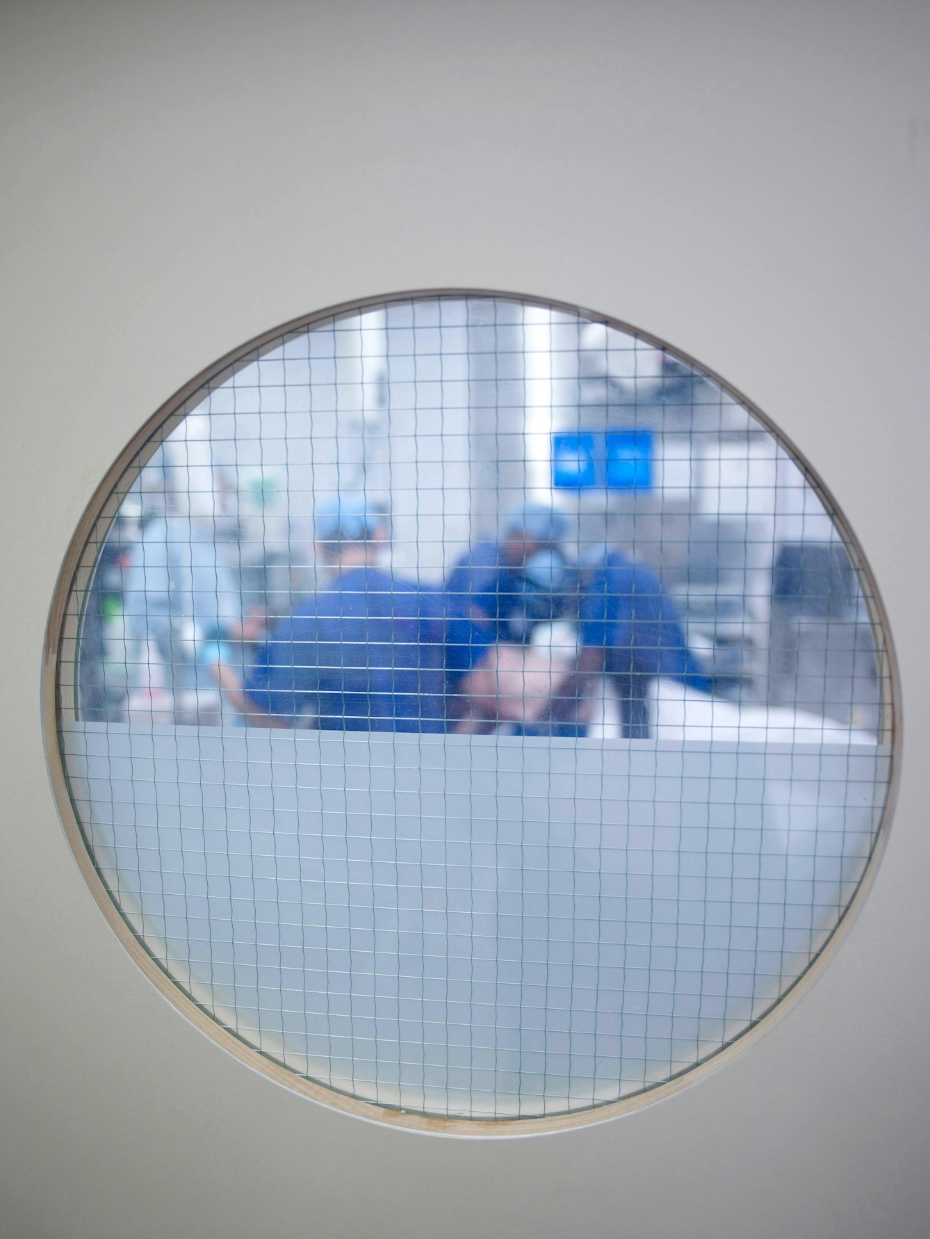 Colour photograph taken through a round window looking into an operating theatre, where staff in blue scrubs, hair covers and masks can be seen preparing the room.