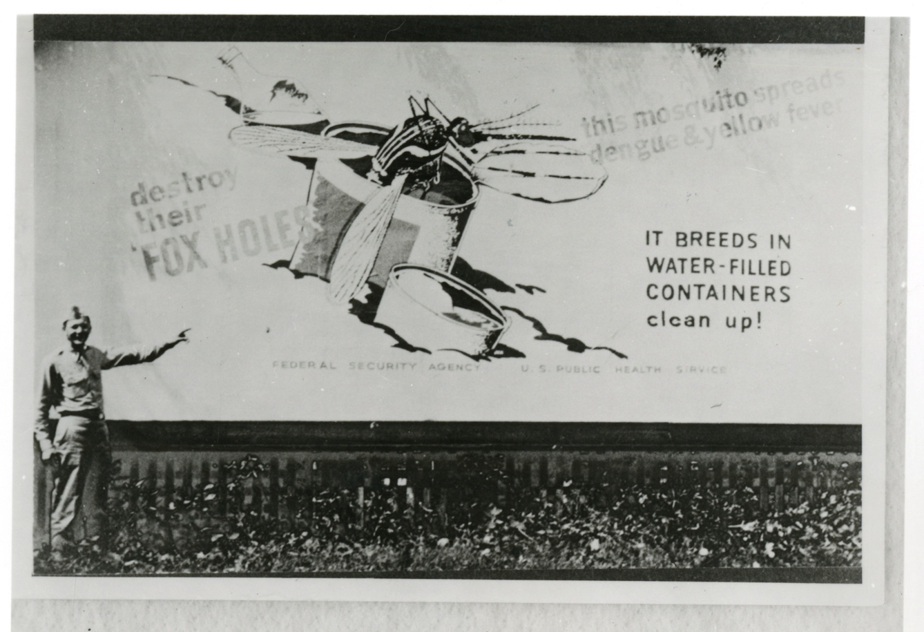 Black and white photograph of a man in military uniform pointing to a billboard on which is the image of a mosquito breeding in a discarded can filled with water.