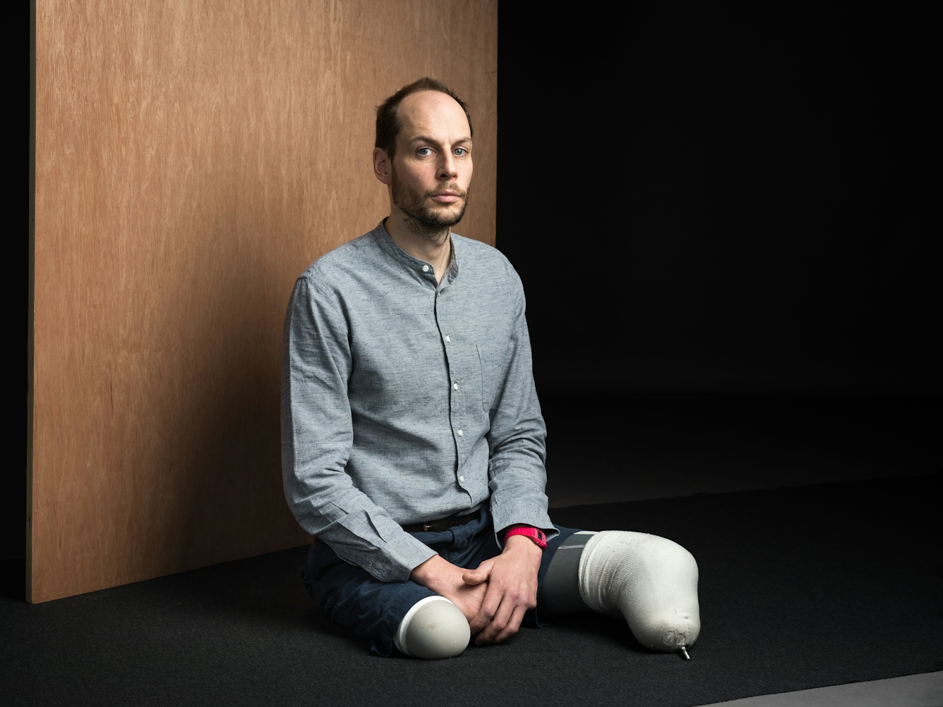 Portrait photograph of Harry Parker. He is wearing a grey shirt and jeans and is sat on a black mat on the floor. He has short brown hair and a beard. There is a wood panel behind him. 