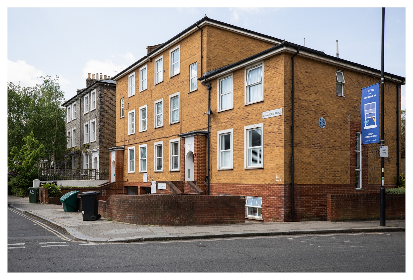 Photograph of a South London street scene in Brixton, showing residential buildings. The main building is a sand brick coloured three story building which is on the corner of two streets. On the side of the building is a circular blue plaque.