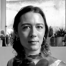 Black and white photographic head and shoulders portrait of Es Morgan who has shoulder-length dark hair and is wearing a patterned top. In the background, there is a window ledge with several small cacti and succulents. 