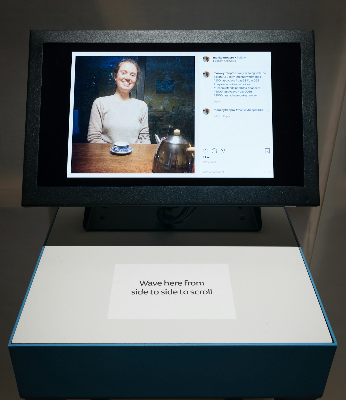 Photograph in an exhibition gallery apace, showing a computer screen mounted on a gallery plinth. On the screen is an image of a young woman seated at a wooden table with a blue china teacup and saucer in front of her and a shiny metal teapot. She is smiling to the camera. To the right of the image is the context graphics of an Instagram comments section include 3 comments. On a label stuck to the plinth in front of the screen are the words, 'Wave here from side to side to scroll'.