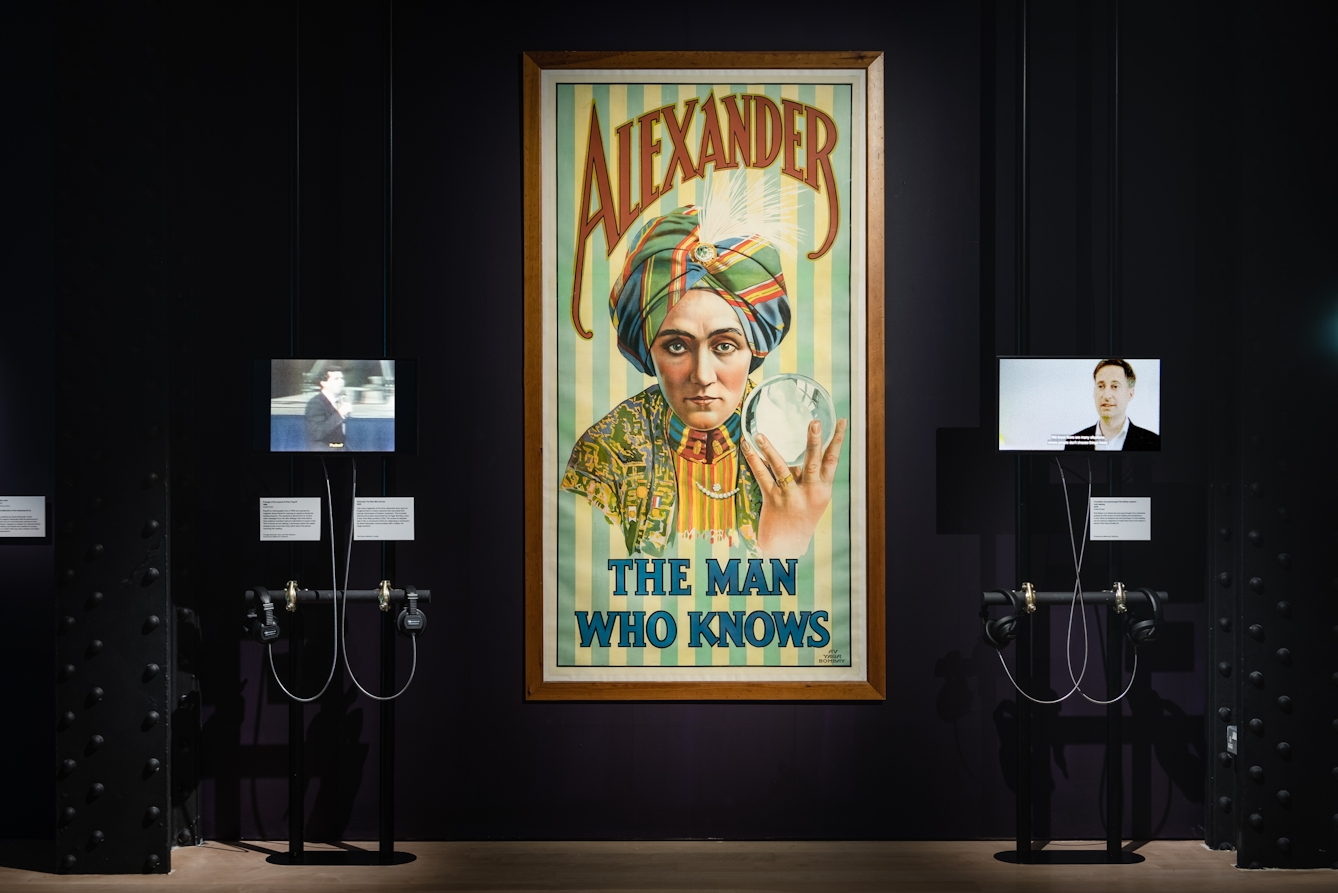 Photograph of a framed poster with text advertising a performance titled 'Alexander: The Man Who Knows', as part of the Smoke and Mirrors exhibition at Wellcome Collection.