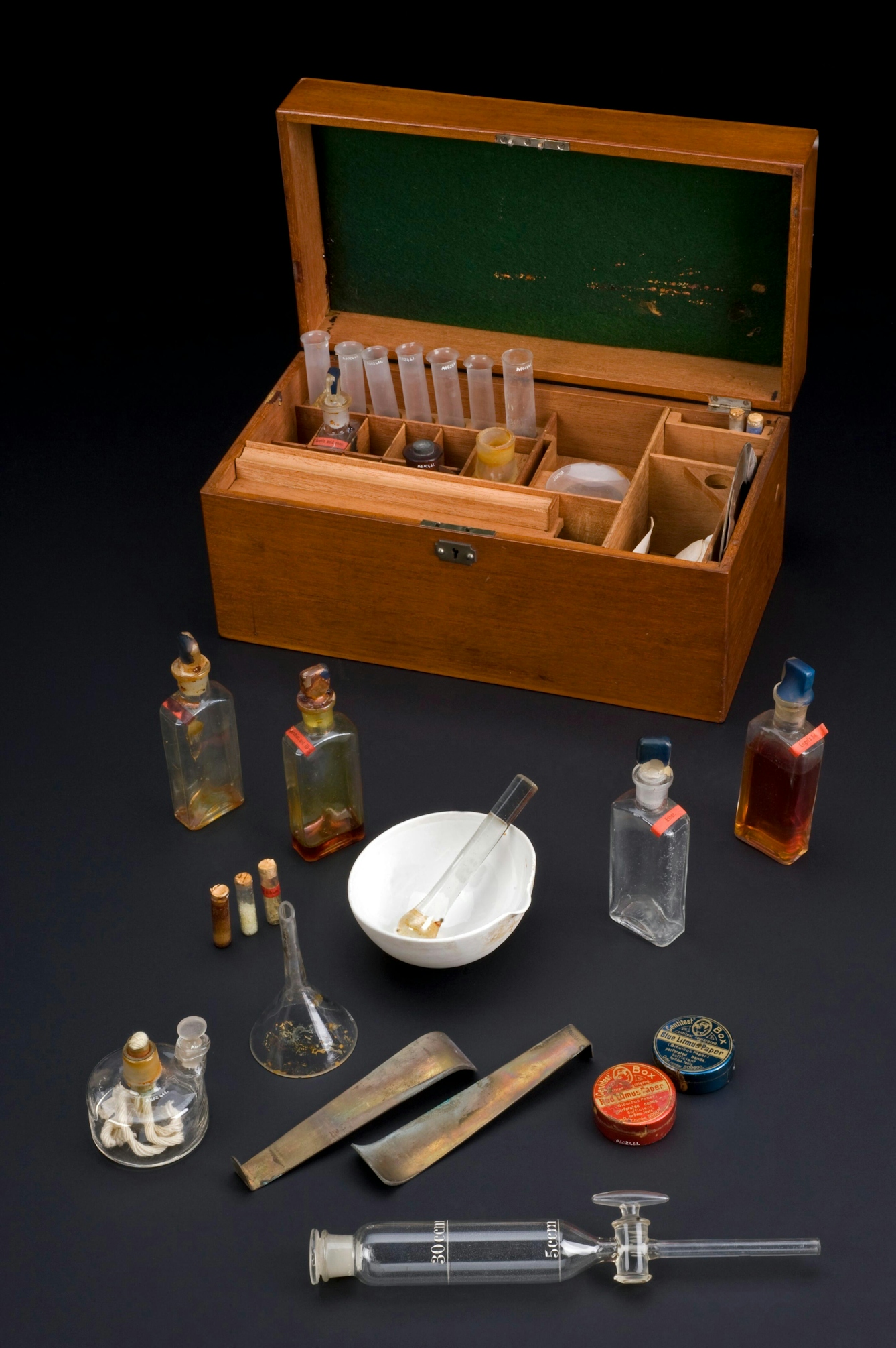 Colour photograph showing a Victorian chemistry set. The wooden storage box is open, and various chemicals and equipment are displayed in front of it, including glass bottles and tubes, round tins, and a mortar and pestle.