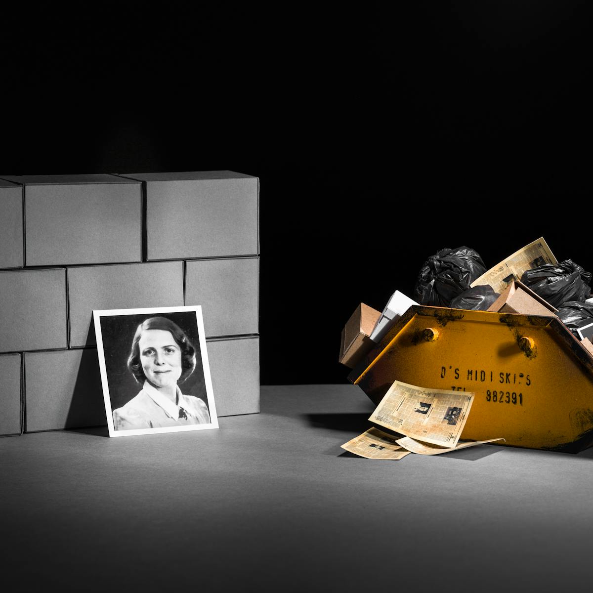 Photograph of a set-built scene made up of a grey card horizontal base against a black vertical background. Built into a mock wall are nine rectangular brick blocks also made out of grey card. To the right of the wall is a yellow skip containing black plastic bags, cardboard boxes and a small photographic print of a protein model. In front of the skip are a pile of yellowed newspapers. Leaning against the brick wall is a photographic portrait of a young woman with short hair wearing a blouse. 
