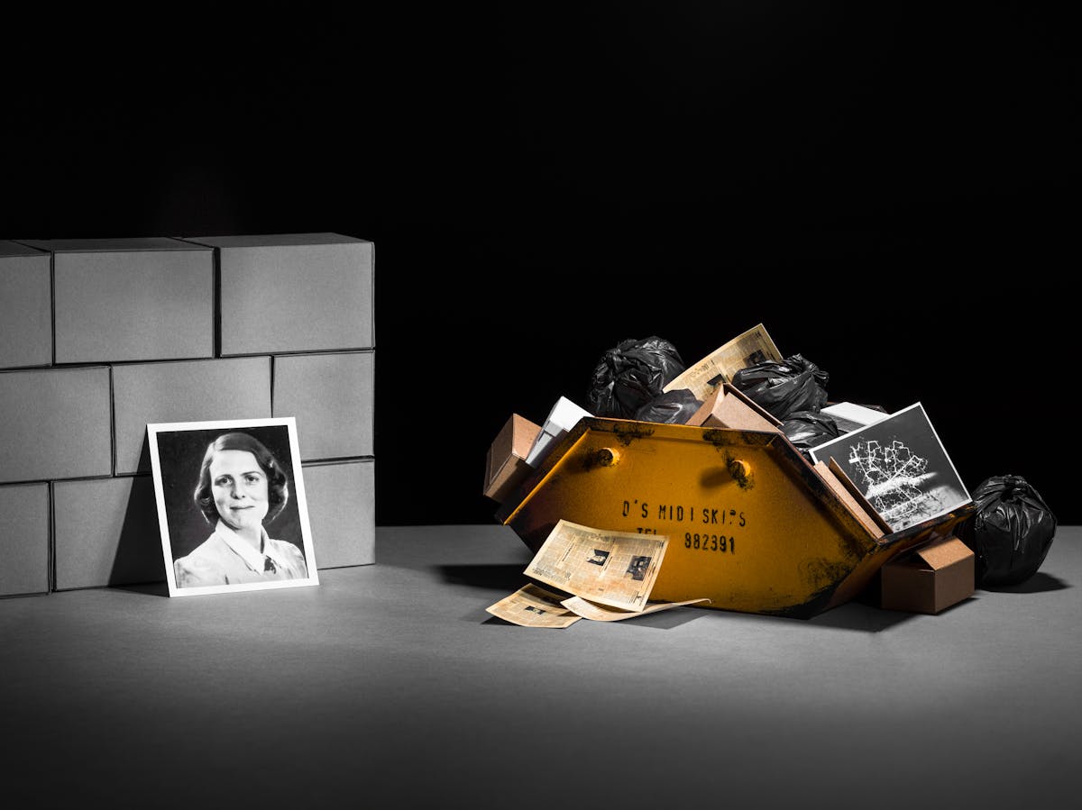 Photograph of a set-built scene made up of a grey card horizontal base against a black vertical background. Built into a mock wall are nine rectangular brick blocks also made out of grey card. To the right of the wall is a yellow skip containing black plastic bags, cardboard boxes and a small photographic print of a protein model. In front of the skip are a pile of yellowed newspapers. Leaning against the brick wall is a photographic portrait of a young woman with short hair wearing a blouse. 
