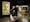 Photograph of a woman in a dark gallery space holding a picture frame in front of her body and part of her face. Within the picture frame you can see straight through her, as if her body and half her face have disappeared. In the background you can see a large historical poster promoting the a magician called "Alexander, the man who knows" and a sawing in half box.