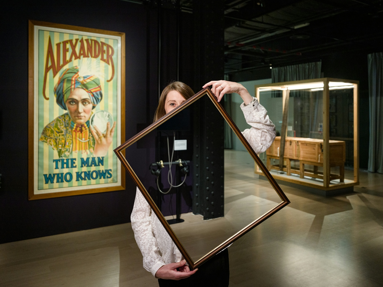 Photograph of a woman in a dark gallery space holding a picture frame in front of her body and part of her face. Within the picture frame you can see straight through her, as if her body and half her face have disappeared. In the background you can see a large historical poster promoting the a magician called "Alexander, the man who knows" and a sawing in half box.