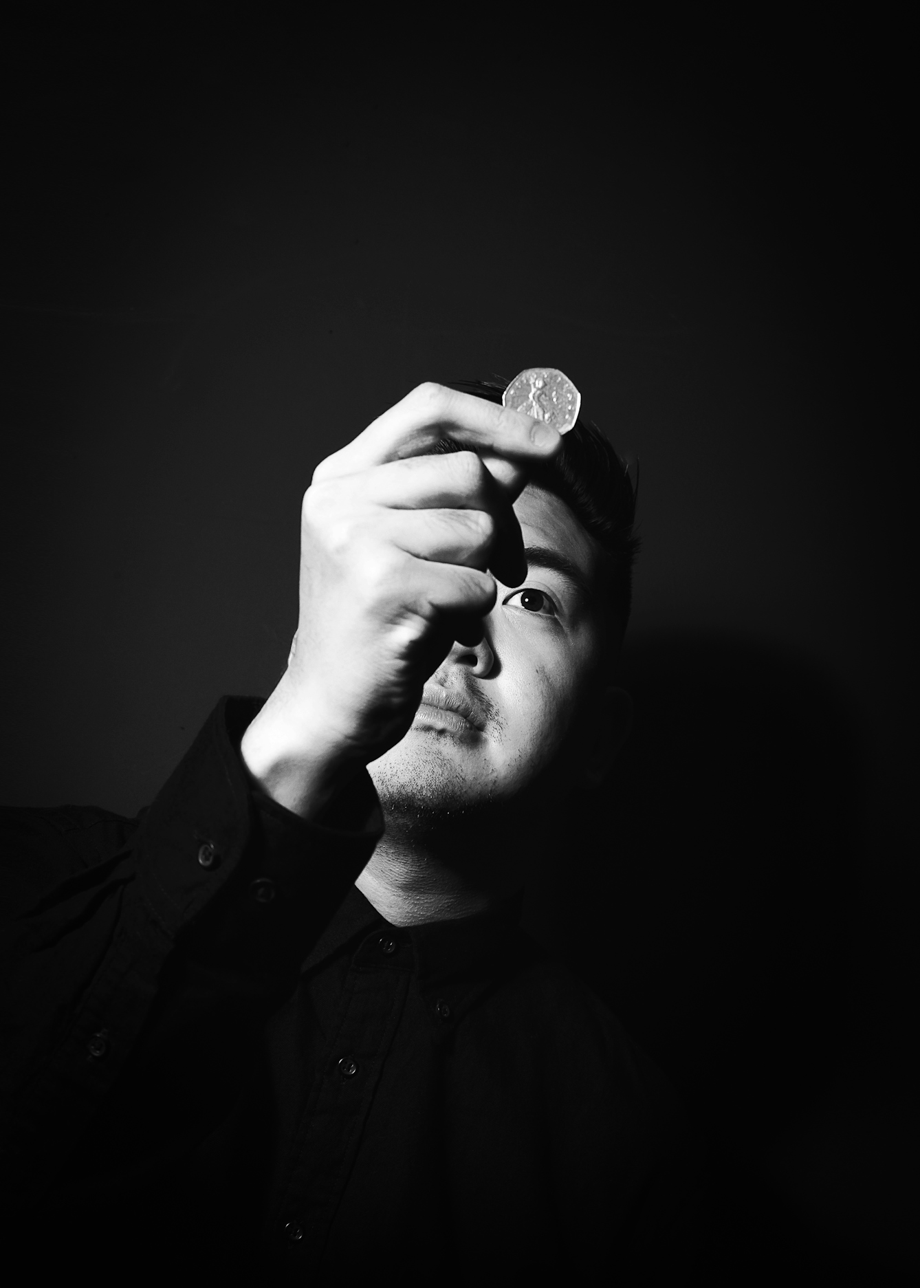 Black and white photographic portrait of a man dressed in a black shirt holding up a 50 pence piece to his face, held in his right hand. He is pictured from the chest up, his eyes directed towards the coin. The man's face and hand is spotlit in a small circle of light. He is standing against a black background which means he is surrounded by darkness.
