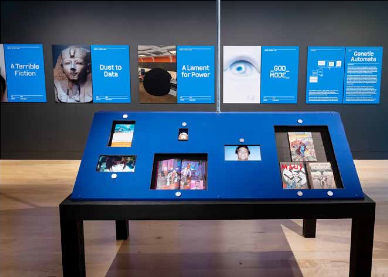 A display case in a museum exhibtion with a sloped blue top showing books, films and videogames.
