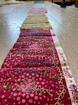 Colour photograph of an embroidery featuring patterned coloured fabric stitched together, stretching into the distance of the image. There are words stitched onto the fabric that are upside-down for the viewer and become unreadable with the distance blur.
