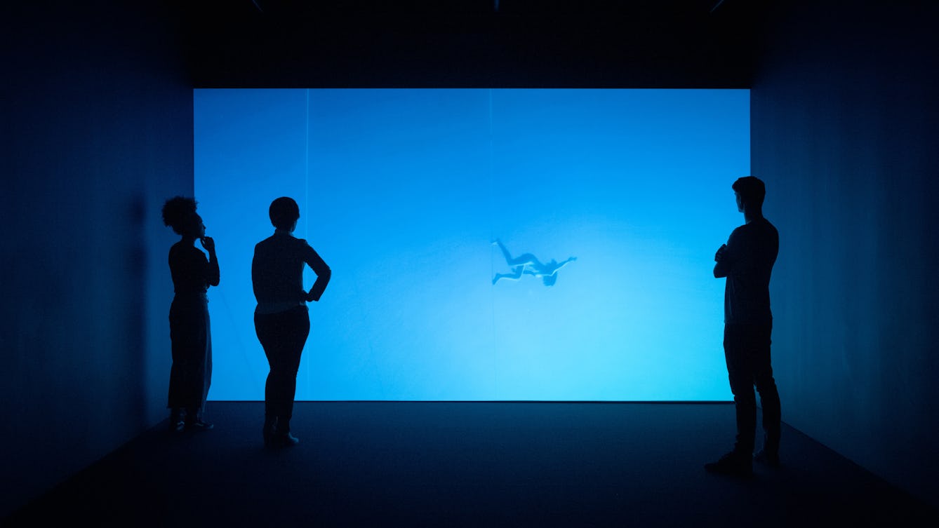 Photograph of visitors exploring a gallery installation. They are standing in the dark in front of a large projection of a figure floating underwater. The room has a blue cast.