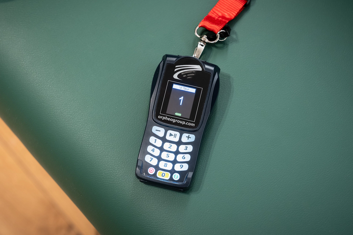 Handheld audio guide device with a keypad and screen, resting on a green bench.