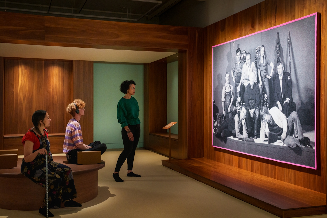Photograph of a gallery exhibition space showing large tapestry hung on a wooden wall. The tapestry is made up of a black and white, archive photograph, from the late 20th century showing a group of white men and women gathered in a group within an exhibition exhibit. Sat crosslegged on a seat in front of the tapestry is a gallery visitor in a striped top. They are listening to the gallery audio guide on a pair of headphones. Either side of them are two more visitors, one standing looking at the tapestry, the other seated holding a walking stick.