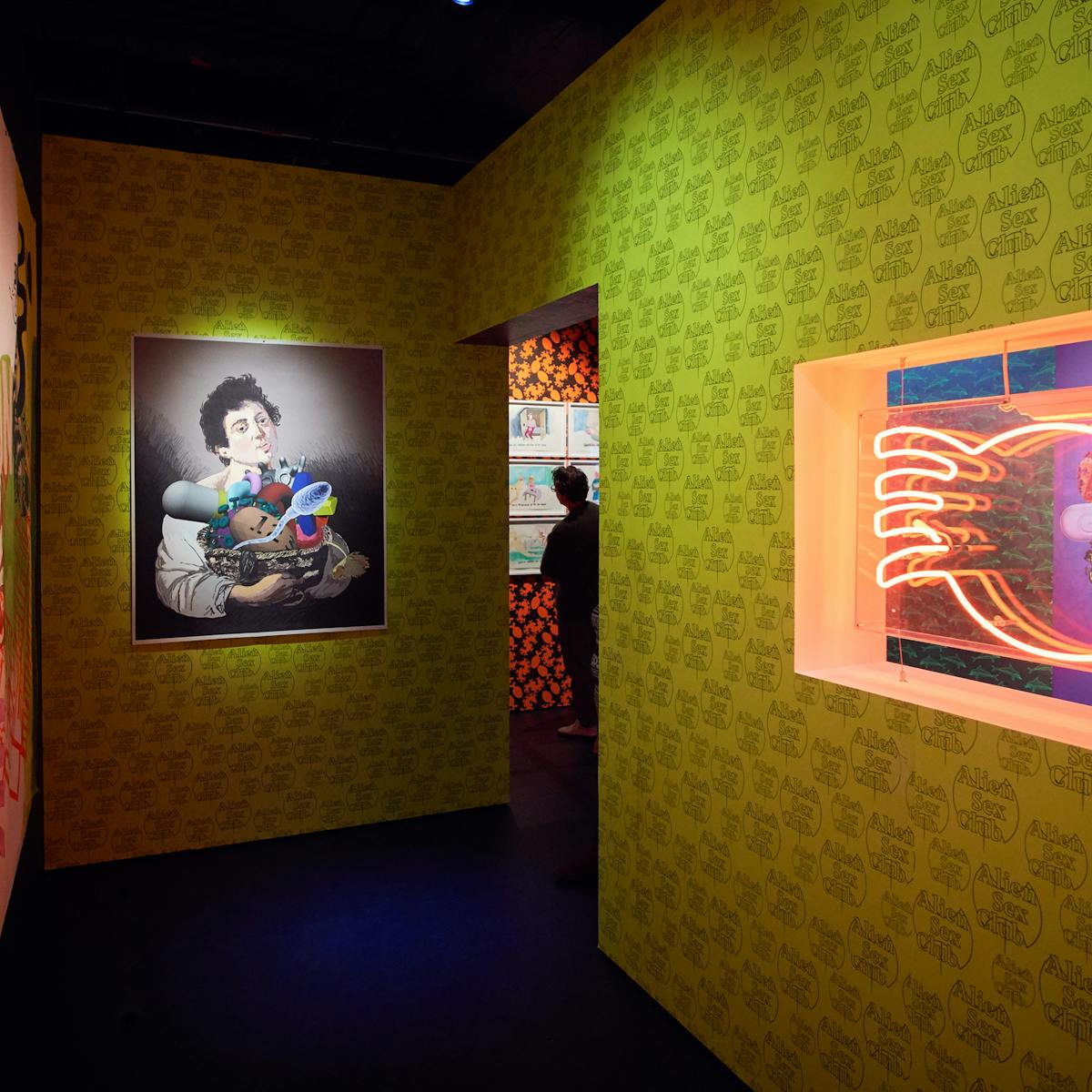 Photograph of a visitor in the distance looking at framed artworks on the wall. In the foreground is colourful patterned wallpaper, a work of art and a red neon exhibit.