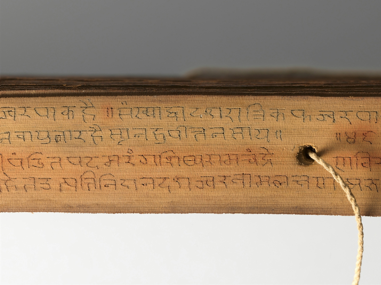 Deail of a strip from a palm leaf manuscript showing text inscribed in Hindi and a string connecting all the leaves emerging from a hole in the strip