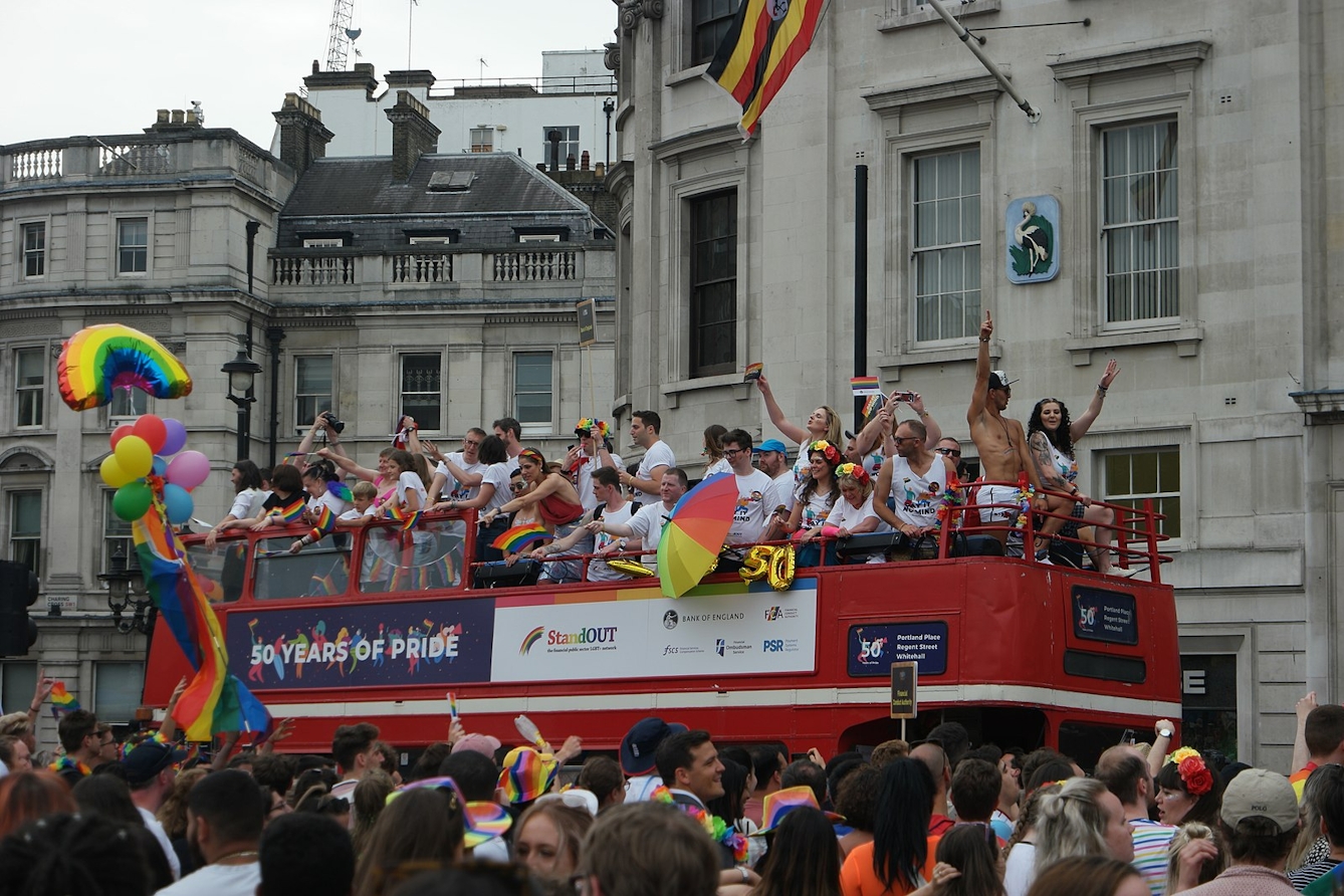 Colour photo of an open top double decker London bus with people waving rainbow flags and celebrating in the street and on the bus