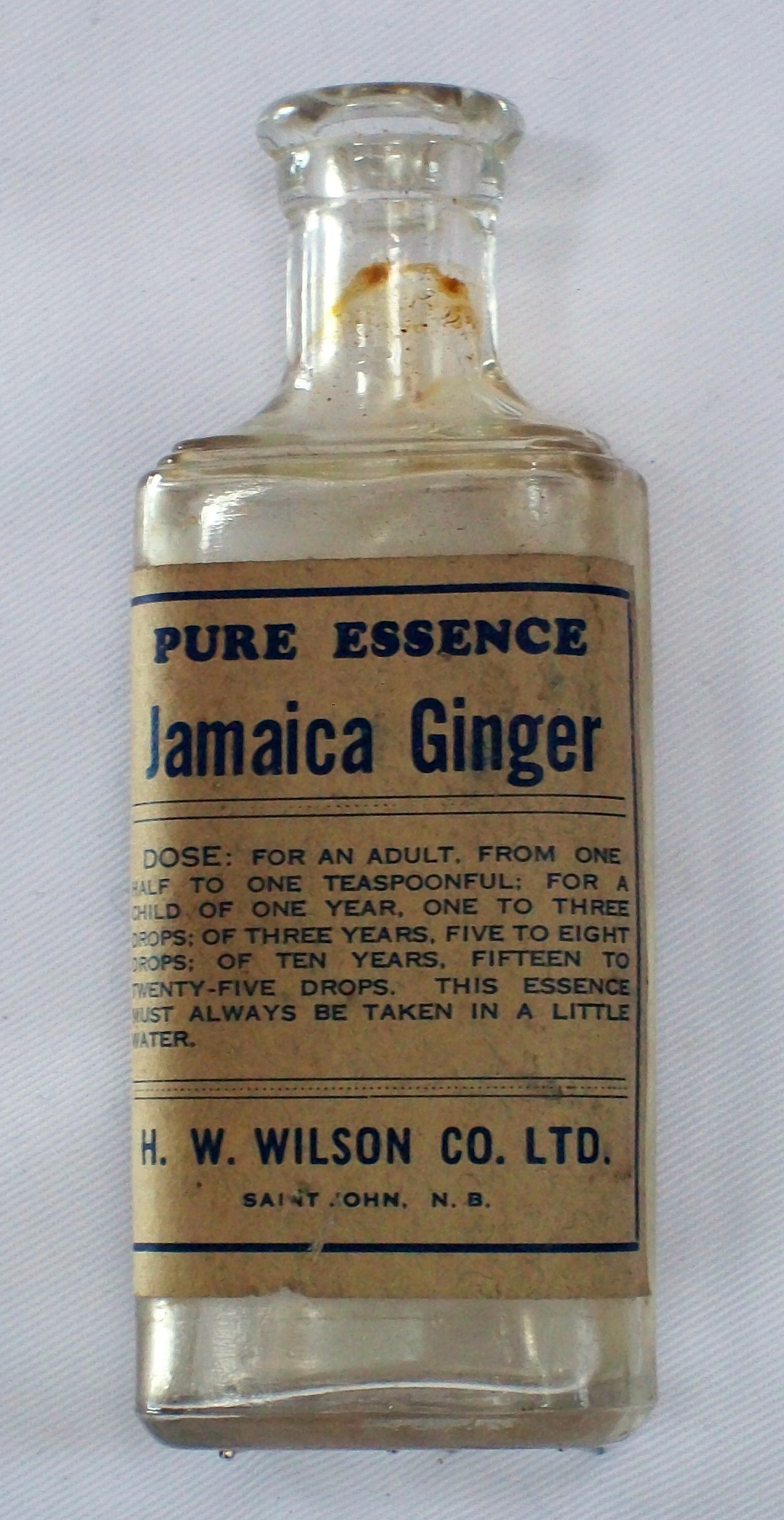 A glass bottle with browned-paper label saying: Pure essence Jamaica Ginger. Recommended dose and manufacturer's details written below.