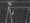 Black and white still showing a man with his hand bandaged up tightly, perched at the top of a ladder about to climb onto a plank balanced between his ladder and another.