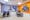 A photograph showing a room containing a desk, computer and two chairs with their backs to the wall. On a lilac painted wall there are three rectangular paintings on canvas boards, of three different sizes, small, medium and large running from left to right. Each painting appears as a collage made up of triangles and quadrilaterals of a various colours floating on different blue backgrounds. The adjacent wall is painted bright orange on which another larger painting, similar in design, hangs. This time the shapes float on a solid grey background.
