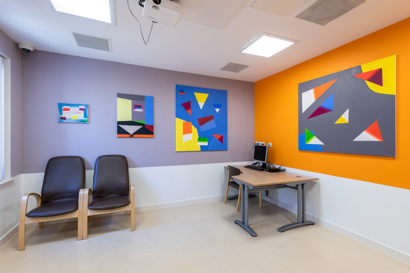 A photograph showing a room containing a desk, computer and two chairs with their backs to the wall. On a lilac painted wall there are three rectangular paintings on canvas boards, of three different sizes, small, medium and large running from left to right. Each painting appears as a collage made up of triangles and quadrilaterals of a various colours floating on different blue backgrounds. The adjacent wall is painted bright orange on which another larger painting, similar in design, hangs. This time the shapes float on a solid grey background.