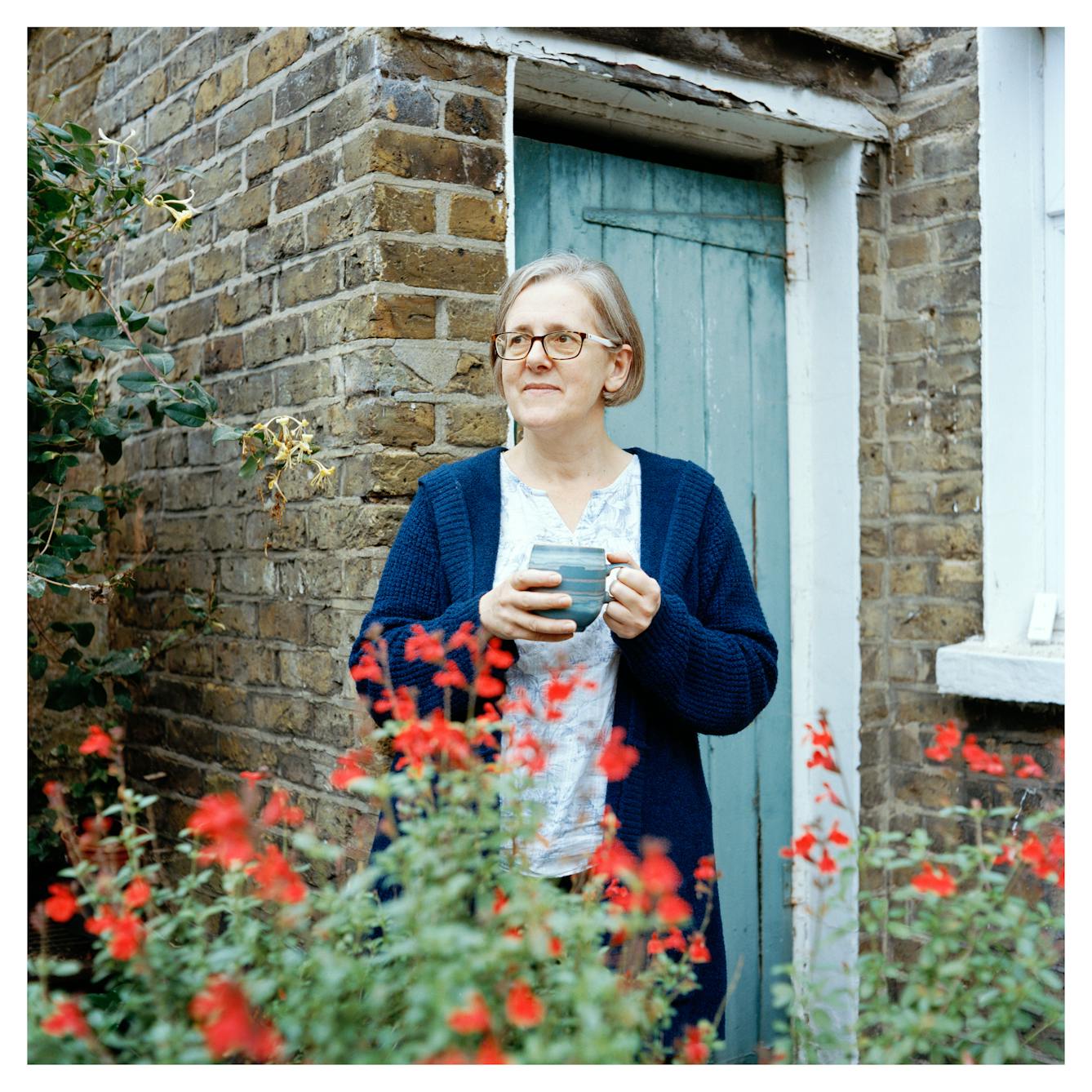 Portrait photograph of Emily, a woman with short grey hair wearing glasses. She is wearing a patterned top and a long blue cardigan and is holding a mug with both hands. 

She is standing in front of a brick building with a blue painted wooden door. She is looking into the distance and in front of her are some red wildflowers. 