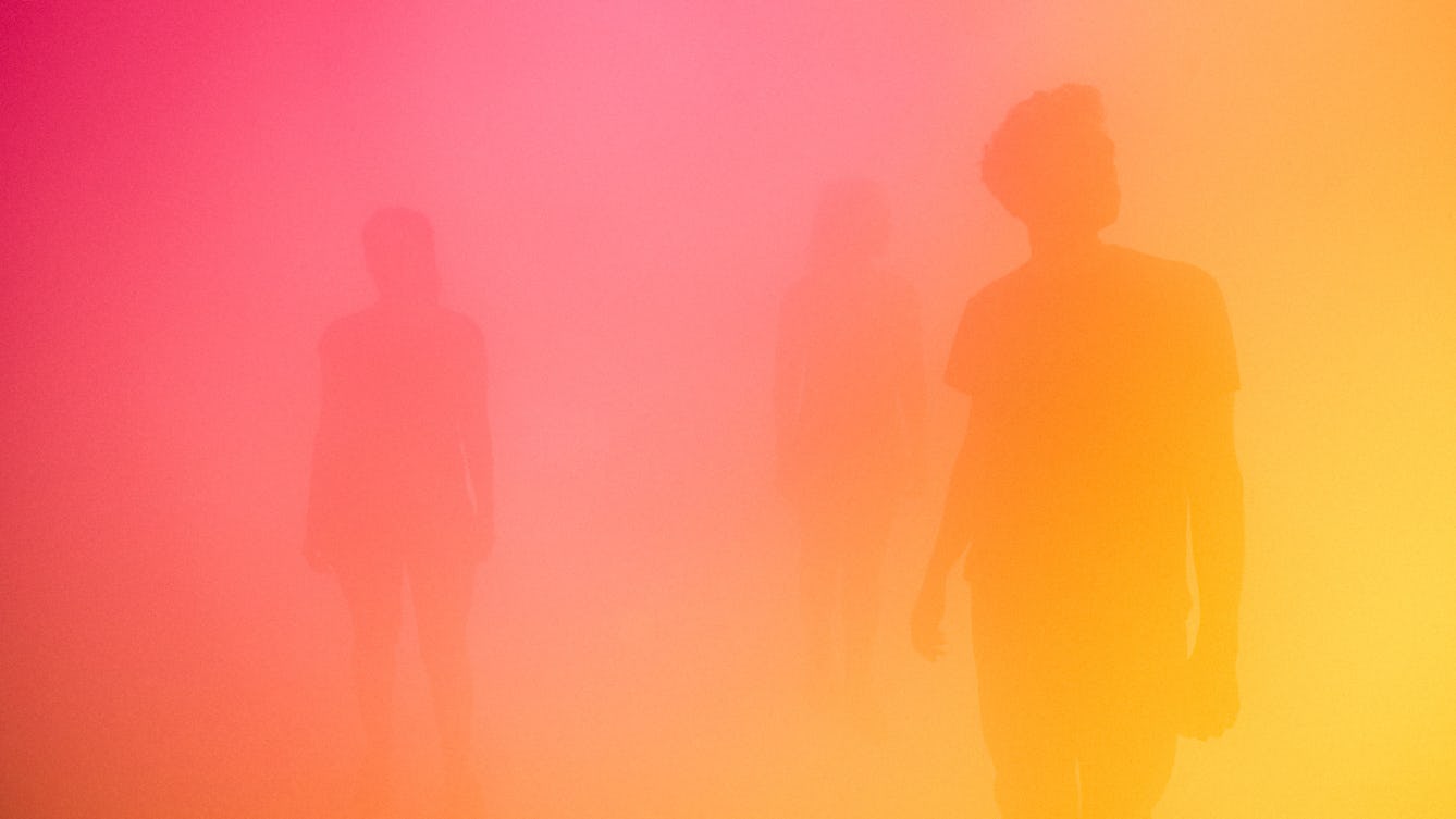 Photograph of three silhouettes in a room full of mist, bathed in a yellow and pink light.