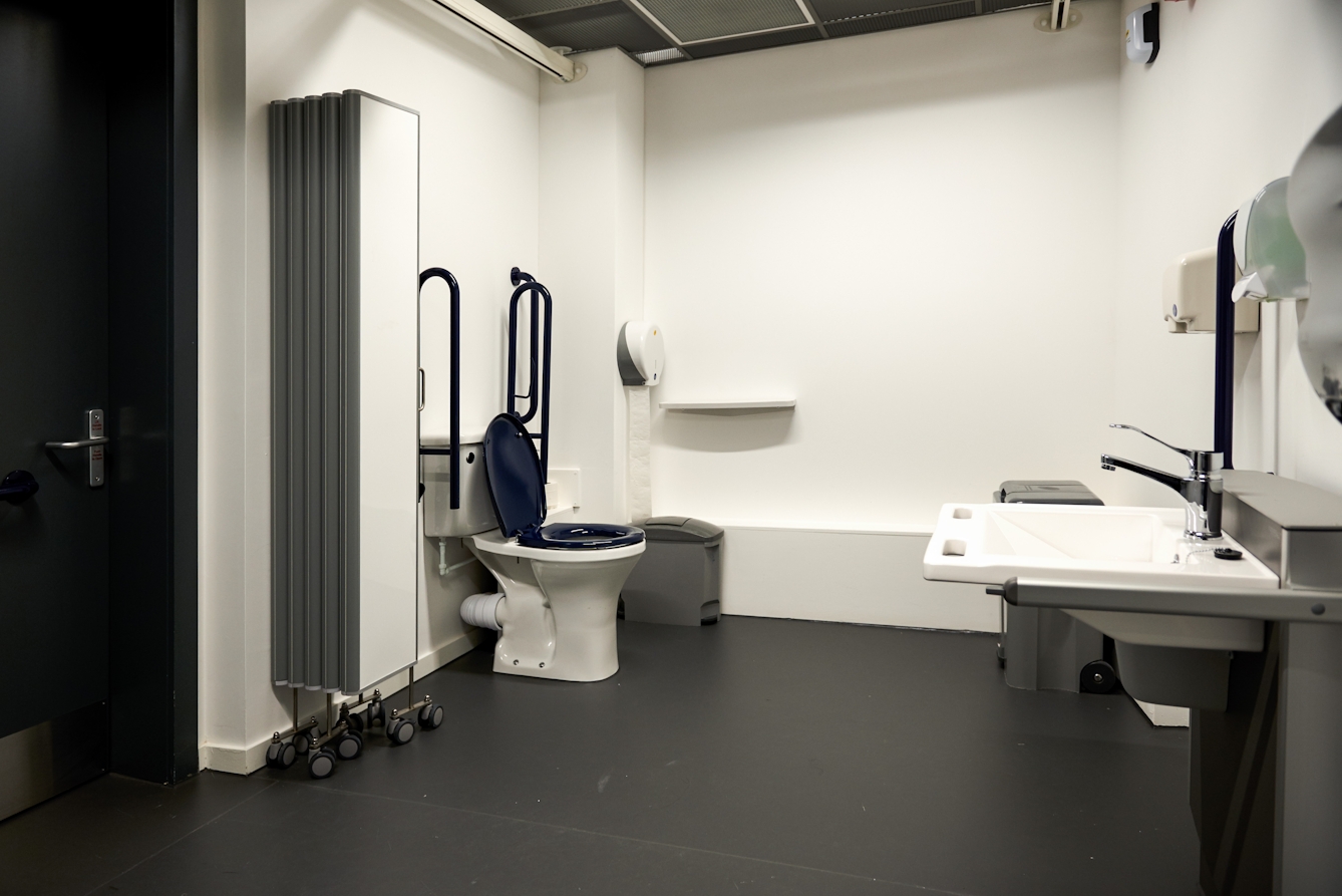 Photograph of the interior of a modern looking accessible toilet, showing the toilet, a screen on wheels, basin and sanitary bins.