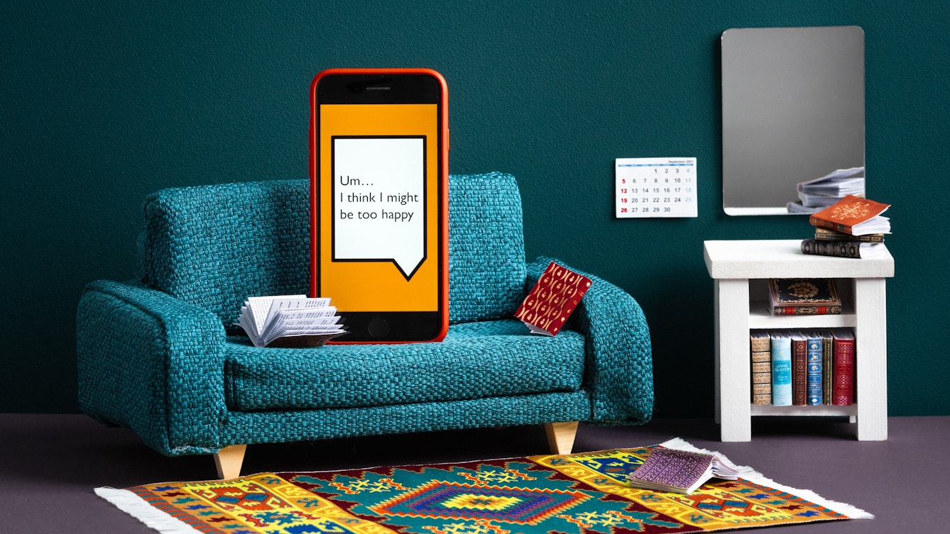 Photograph of a set built scene using miniature dolls house furniture. The scene shows a blue textured fabric sofa on which a smartphone in an orange case rests vertically on the cushion as if sitting up. On the floor in front of the sofa is a colourful aztec patterned rug on a moave coloured floor. To the right of the sofa is a white side table containing tiny books on its lower shelf and a stack of books on its tabletop. Hung on the blue painted wall behind the sofa and table are a mirror and month view paper calendar. Surrounding the smartphone are a couple more books, one of them lying open. On the screen of the phone is a large white angular speech bubble, outlined with a black line, against an orange background. Within the speech bubble are the words "Um... I think I might be too happy".