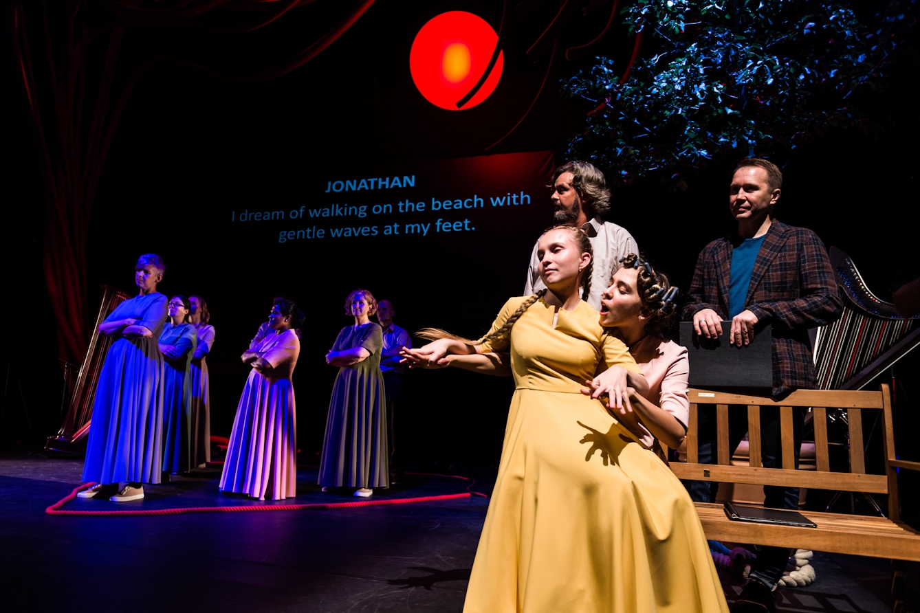 Photograph of a theatre stage. In the foreground and lit by stage light, a female actor wearing a mustard yellow formal costume is sitting on the knee of another actor. The actor in yellow is resting her arms and palms over the arms and hands of her fellow actor, who is speaking over her shoulder. In the background a number of other actors standing in a triangular formation within the confines of a red rope laying on the floor. On the projection in the far background, blue text reads “Jonathan: I dream of walking on the beach with gentle waves at my feet.”