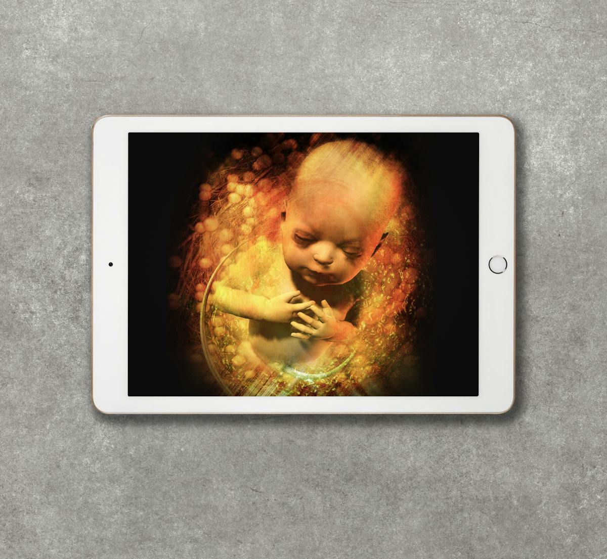 Photograph of a white tablet device resting on a grey concrete textured background. On the screen is a colour illustration of a baby in the womb. The head, shoulders and chest of the baby is visible, and it is surrounded by swirling abstract patterns. The baby has its eyes closed and its arms crossed in front of it, with its hands resting lightly on its chest. 