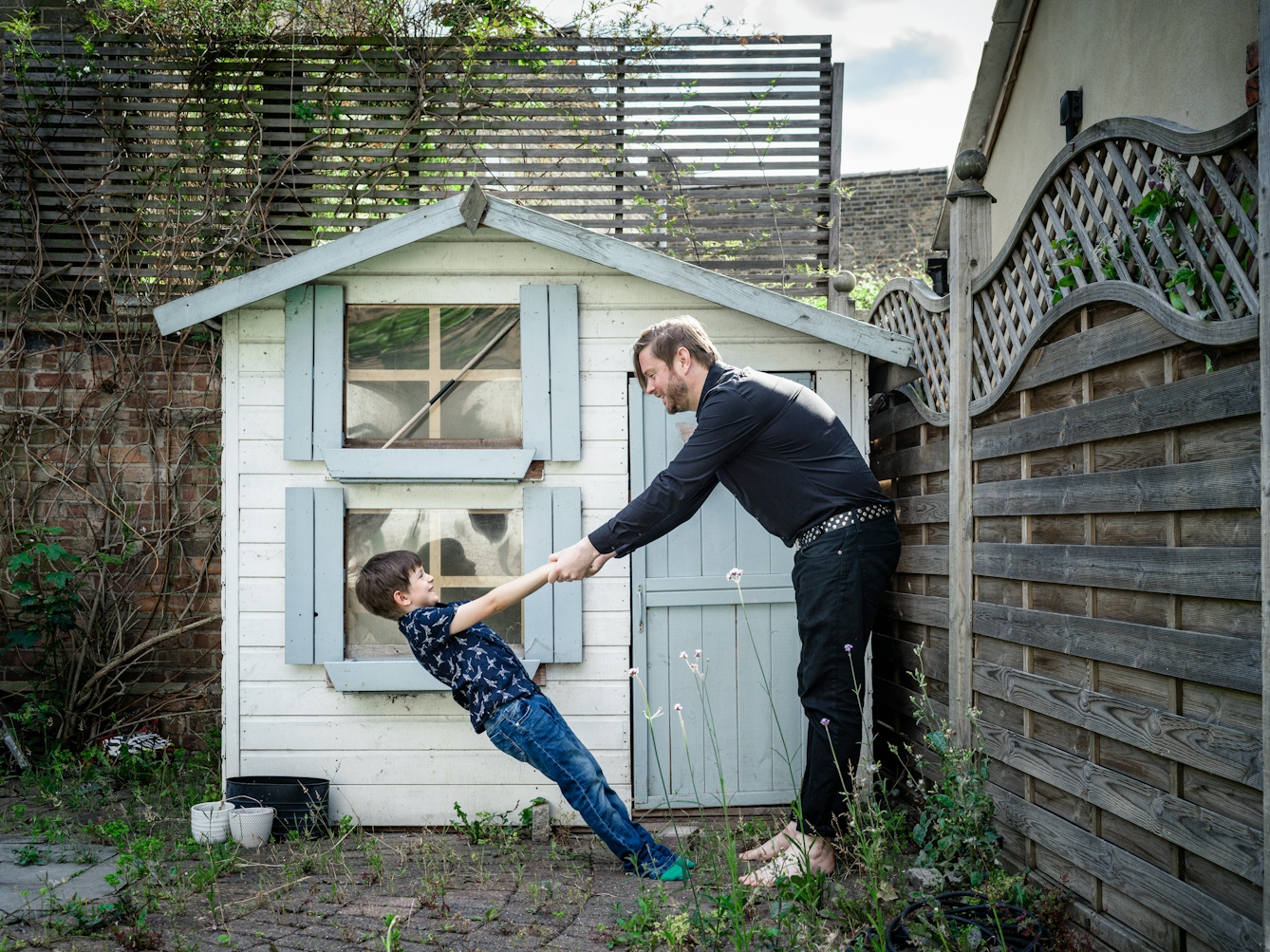 Photograph of a man and his young son in their back garden in front of a Wendy house. They are holding hands and the young son is leaning back at an extreme angle, held up only by his father.
