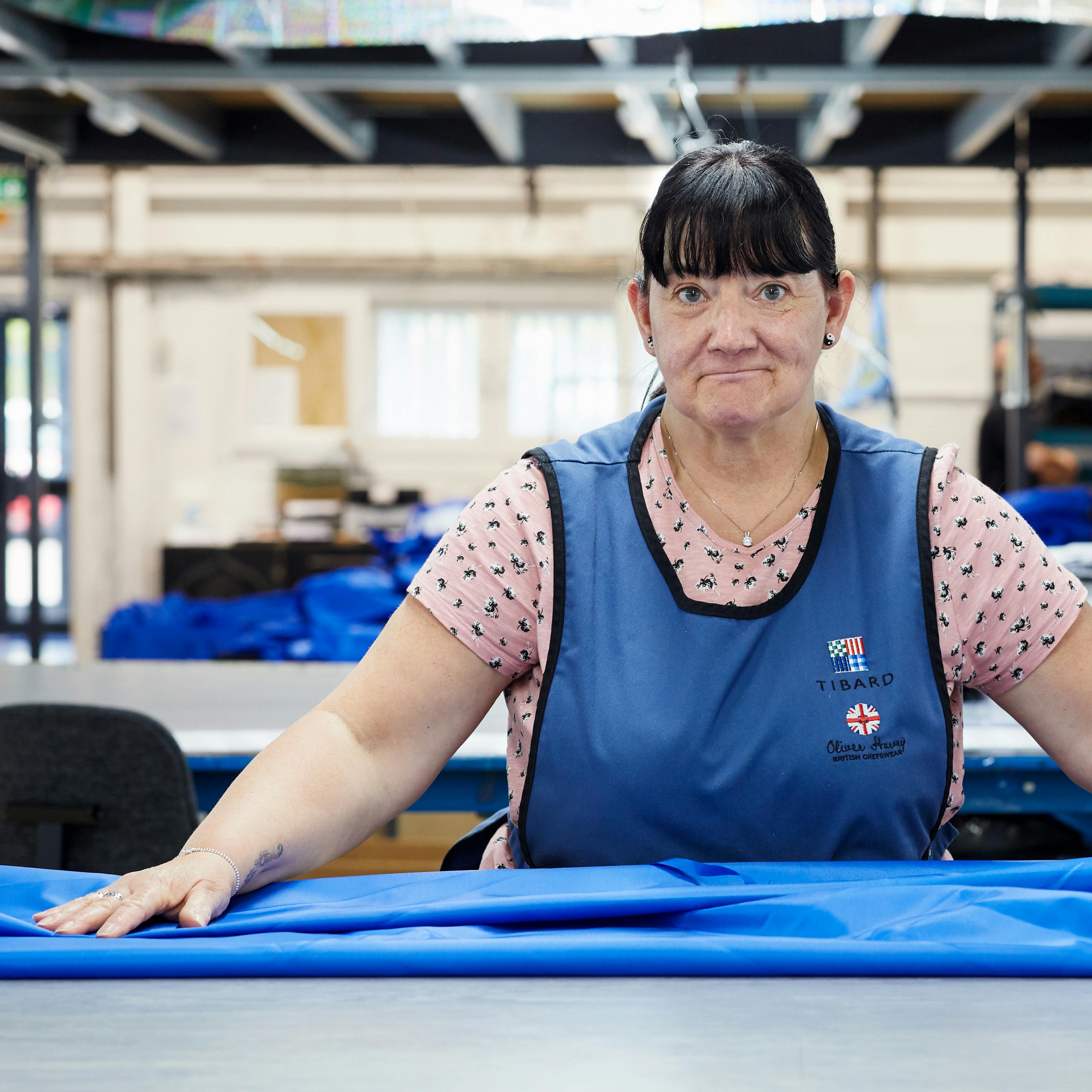 Photograph of a woman in a textile factory wearing a blue tunic. She is look straight to camera with her hands outstretched to either side resting on a blue medical garment. In the background are cardboard boxes and other workstations.