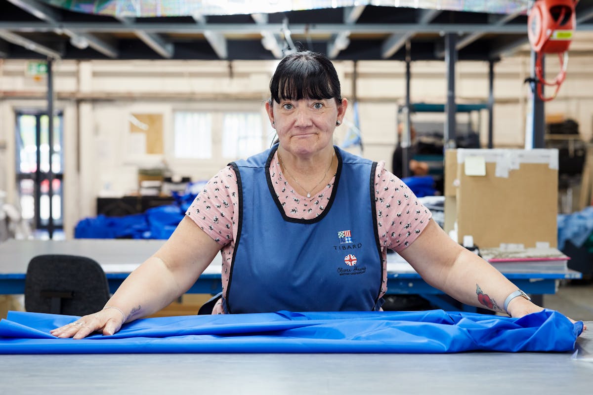 Photograph of a woman in a textile factory wearing  a blue tunic. She is look straight to camera with her hands outstretched to either side resting on a blue medical garment. In the background are cardboard boxes and other workstations.